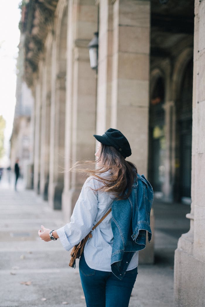 A stylish woman walks along a historic urban street, wearing denim and a black hat.