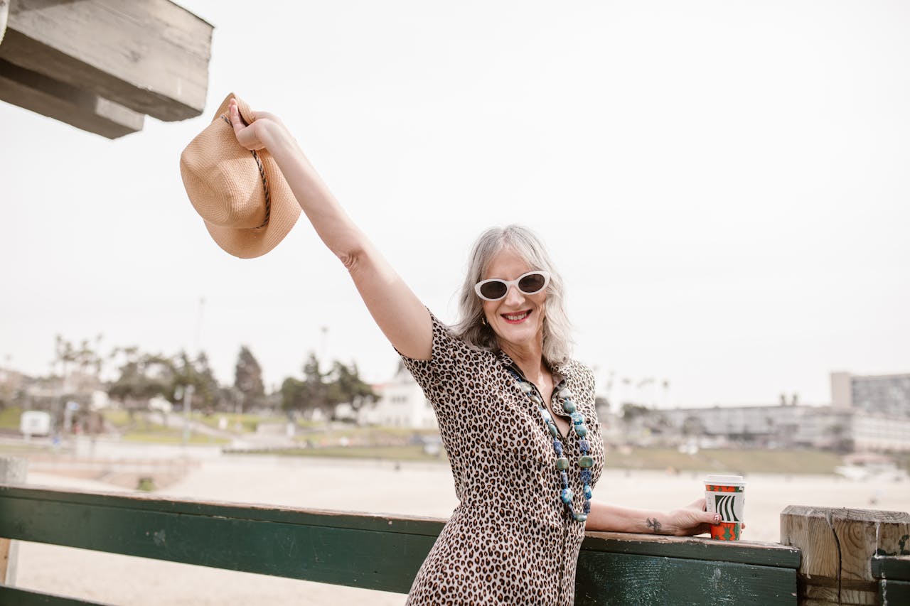 heros-img Stylish senior woman with hat at the beach, embracing life joyfully.