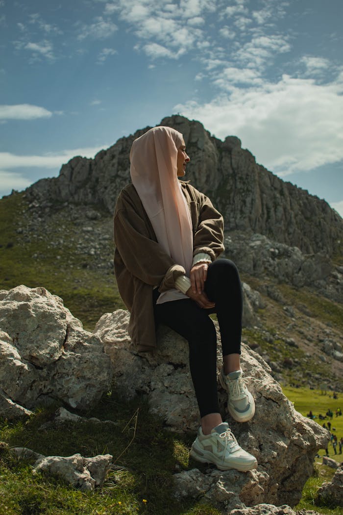 A woman in a hijab sits on a rocky mountaintop, embodying serenity and nature.